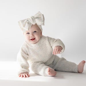 Könnte beinhalten: Ein cremefarbenes Strickset bestehend aus einem Pullover und einer passenden Hose mit einem dezenten, mehrfarbigen Sprenkelmuster. Das Outfit wird durch ein großes, passendes Strick-Stirnband mit Schleife und demselben Sprenkelmuster ergänzt.