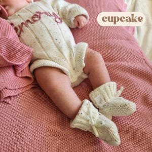 May include: A cream-colored knit romper with multicolored flecks and the word "memory" embroidered in dark pink thread across the chest. The romper is paired with matching cream-colored knit booties that feature a small white tag with "cgl" text.