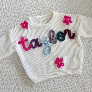 May include: White knitted sweater with the name "Taylor" spelled out in colorful yarn and pink flower embellishments.