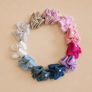 May include: A circular arrangement of multiple pairs of knitted baby booties in a spectrum of colors including beige, pink, magenta, purple, blue, teal, and white. Each pair features a ribbed cuff, a small white brand tag, and matching ties.