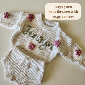May include: A cream-colored knit sweater and matching shorts set featuring multicolored flecks. The sweater displays the name JENSYN embroidered in sage green yarn, accented with rose-colored flowers with sage centers. Text reads: sage yarn rose flowers with sage centers.