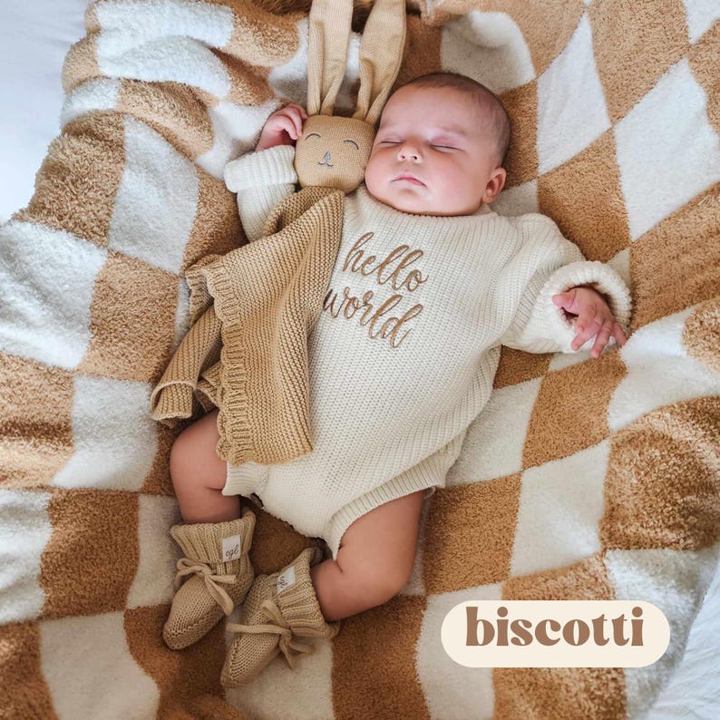 May include: A sleeping infant wearing a cream-colored knit romper with the embroidered text hello world, paired with matching tan knit booties. A tan plush rabbit security blanket rests beside the infant on a tan and white checkered blanket.