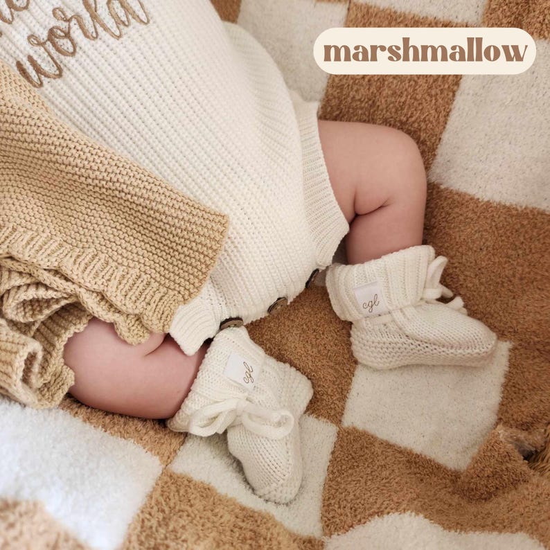 May include: A cream-colored knitted baby romper with brown buttons and matching cream-colored knitted booties featuring white labels with the letters cgl. The romper has a ribbed texture and is paired with a tan knitted blanket.