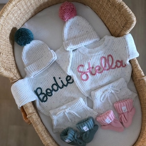 May include: Two sets of white speckled knit baby clothing, each including a long-sleeve sweater with a name embroidered in dark green or pink, a matching beanie with a pom-pom, knit shorts with a drawstring, and a pair of small knit booties.