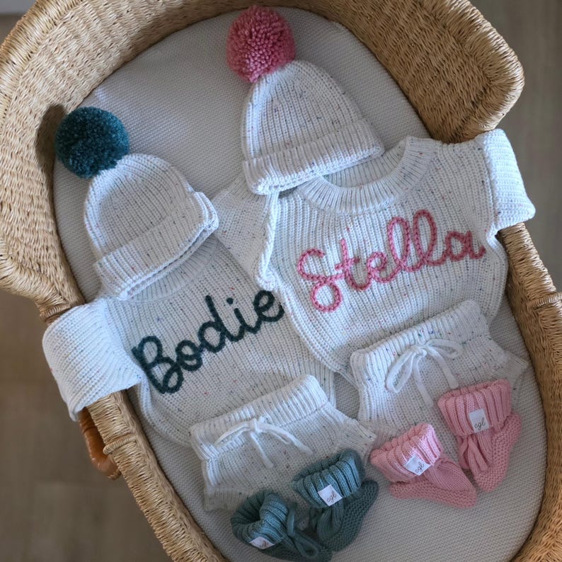 May include: Two sets of white speckled knit baby clothing, each including a long-sleeve sweater with a name embroidered in dark green or pink, a matching beanie with a pom-pom, knit shorts with a drawstring, and a pair of small knit booties.