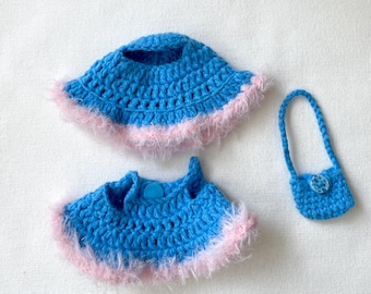 May include: A set of handmade crochet doll clothes in bright blue with a pink fluffy trim. The set includes a hat, a top, and a small purse with a button closure. The items are displayed on a white surface.