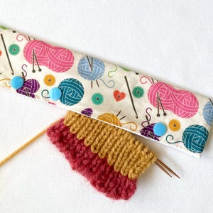 May include: A cream-coloured knitting needle case with a colourful pattern of yarn balls, needles, buttons, and hearts. The case is closed with three light blue buttons. Below the case is a knitted swatch in shades of red and gold, with two knitting needles.