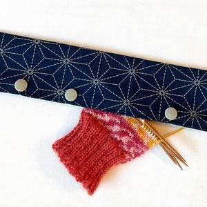 May include: A rectangular dark blue fabric case with a white geometric star pattern and three light grey snap buttons. A partially knitted sock in shades of red, pink, and orange is tucked into the case with several thin metal knitting needles.