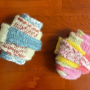 May include: Two hand-knitted leg warmers featuring a textured, ribbed stitch pattern. The left warmer displays a colour palette of light blue, cream, and speckled brown. The right warmer features a vibrant mix of pink, yellow, and cream yarn.
