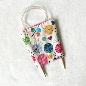 May include: A fabric needle case with a white background featuring a colourful pattern of yarn balls, buttons, and hearts. The case is secured with a bright blue snap button and holds circular knitting needles with a clear plastic cord.