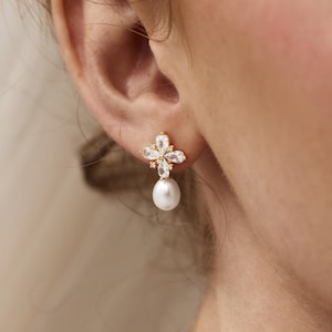 Peut inclure: Une boucle d'oreille dorée avec un motif floral et une perle pendante. La boucle d'oreille présente un groupe de cristaux transparents disposés en forme de fleur, avec une seule perle blanche et brillante suspendue en dessous. La boucle d'oreille est portée à l'oreille.