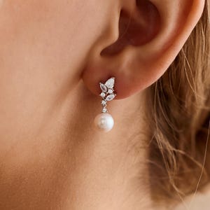 May include: A delicate earring featuring a cluster of small, clear gemstones and a dangling white pearl. The earring is attached to the ear. The design is elegant and suitable for special occasions.