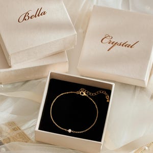 May include: A delicate gold chain bracelet featuring a single small white pearl, presented in an open square cream-colored gift box with a black interior. Two additional closed cream gift boxes are nearby, one labeled Bella and one labeled Crystal.