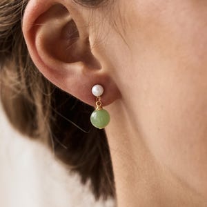 May include: Gold-toned earrings featuring a small white pearl stud and a round, light green gemstone drop. The earrings are worn on an ear, with the focus on the jewelry's design and color.
