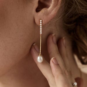 May include: A gold earring featuring a dangling white pearl and a line of small, clear stones. The earring is worn by a person with fair skin. A matching ring with two pearls is also visible.