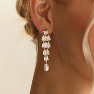 May include: A gold-toned drop earring featuring four tiered rows of clear baguette-cut crystals arranged in a fan-like structure, finished with a single white teardrop-shaped pearl dangling from the bottom of the crystal arrangement.