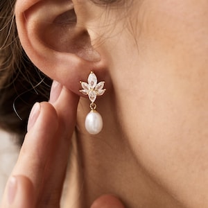 Pode incluir: Um brinco dourado com um design floral e uma pérola pendurada. O brinco apresenta um aglomerado de cristais transparentes em forma de flor, com uma única pérola branca suspensa abaixo. O brinco é usado em uma orelha.