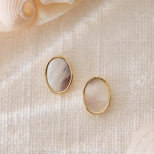 May include: A pair of oval-shaped earrings with a gold rim and a white mother-of-pearl center. The earrings are displayed on a white fabric background.
