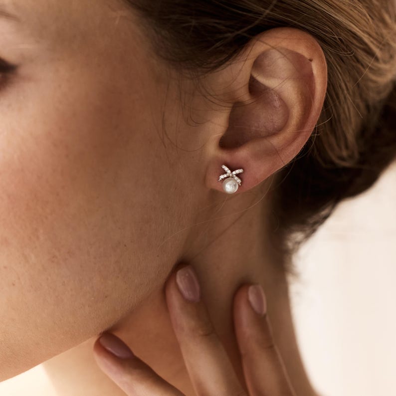 Puede incluir: Primer plano de un pendiente de perla con dise&ntilde;o de lazo. El lazo est&aacute; incrustado con peque&ntilde;os cristales brillantes. El pendiente se muestra sobre el fondo de una oreja y cuello de una persona.