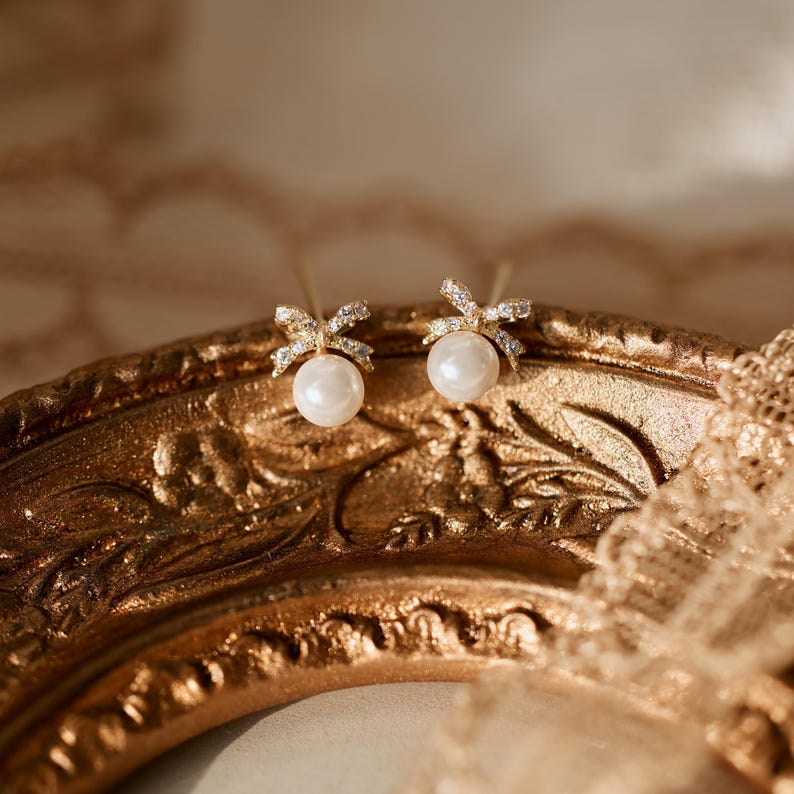 Puede incluir: Un par de pendientes dorados con forma de lazo, cada uno con una perla en el centro. Los lazos est&aacute;n incrustados con peque&ntilde;os cristales transparentes. Los pendientes se exhiben en una bandeja dorada ornamentada.
