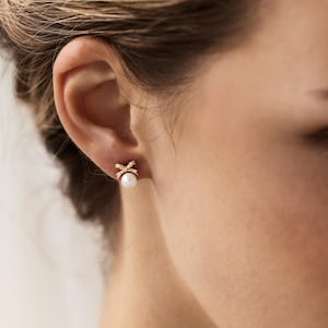 Puede incluir: Primer plano de un pendiente de oro con dise&ntilde;o de lazo y una perla blanca. El lazo est&aacute; incrustado con peque&ntilde;os cristales transparentes. El pendiente se lleva en la oreja de una persona.