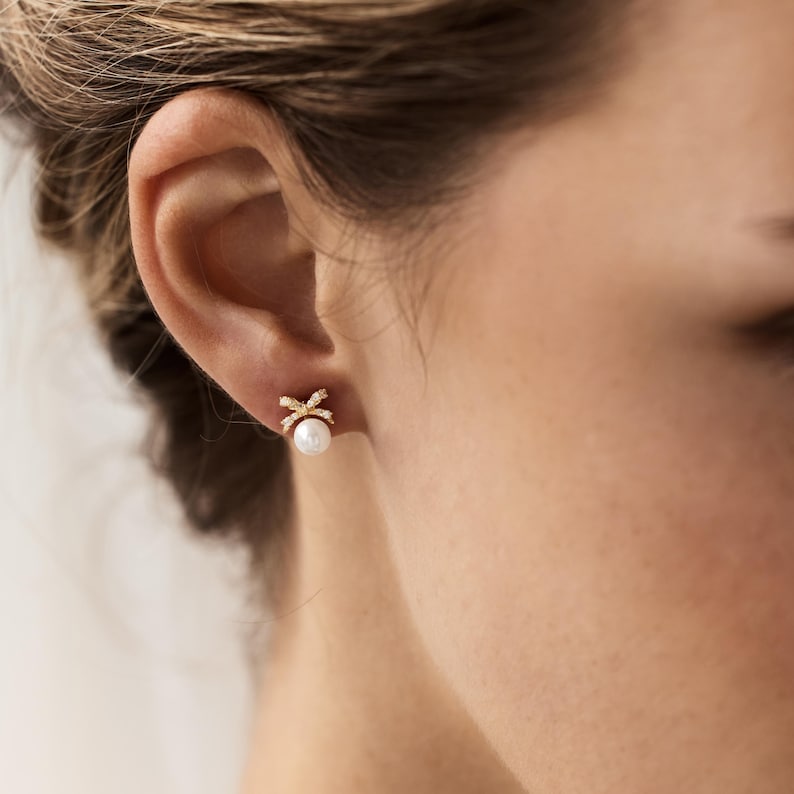 Puede incluir: Primer plano de un pendiente de oro con dise&ntilde;o de lazo y una perla blanca. El lazo est&aacute; incrustado con peque&ntilde;os cristales transparentes. El pendiente se lleva en la oreja de una persona.