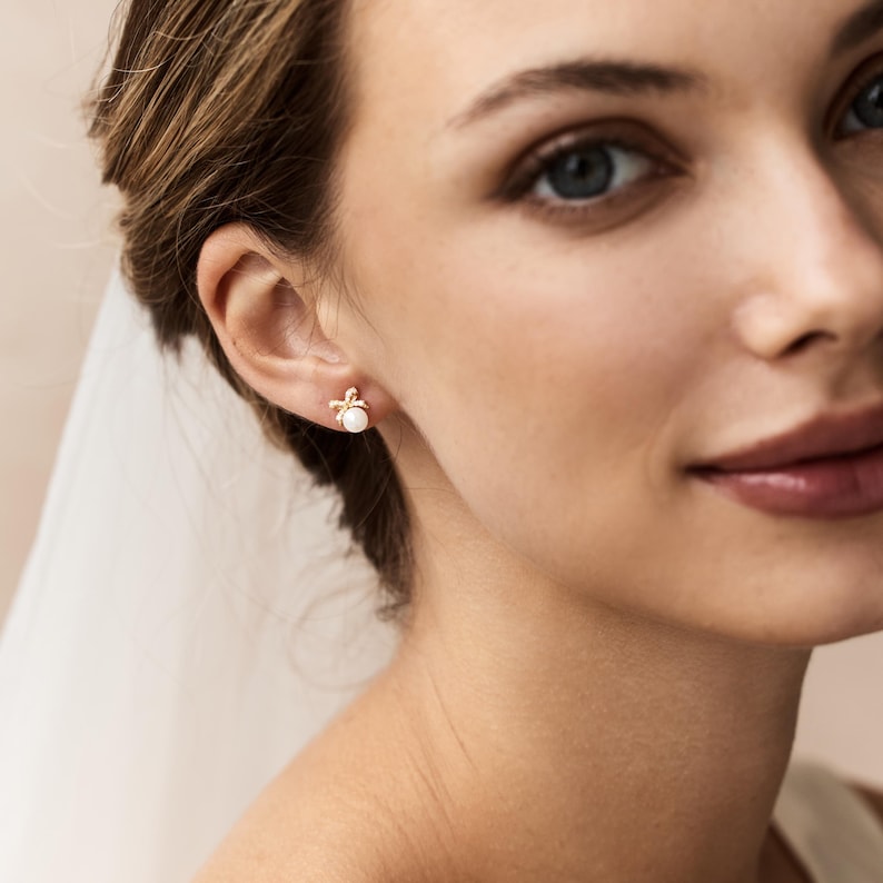 Puede incluir: Primer plano de una mujer con pendientes de oro. Cada pendiente presenta una peque&ntilde;a perla blanca redonda en el centro, rodeada por un delicado dise&ntilde;o dorado. Los pendientes destacan sobre la piel de la mujer, resaltando su elegancia.