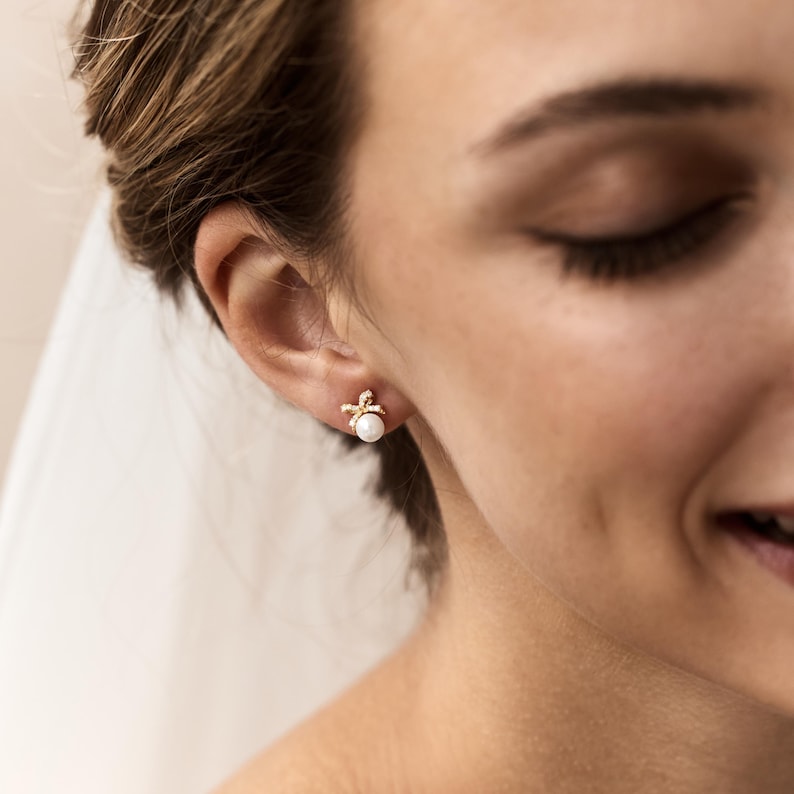 Puede incluir: Primer plano de la oreja de una mujer adornada con un pendiente de oro. El pendiente presenta una peque&ntilde;a perla redonda en el centro, rodeada de un delicado dise&ntilde;o dorado con peque&ntilde;os acentos brillantes. La mujer sonr&iacute;e.