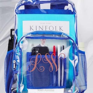 May include: Clear backpack with blue accents, filled with books and stationery. Features a front pocket with a monogram and a side mesh pocket. Includes a copy of "KINfolk" magazine and a book titled "Mary Poppins".