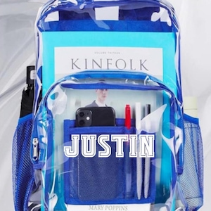 May include: A clear backpack with blue trim and accents. The backpack is filled with books, a phone, and pens. The name "JUSTIN" is printed on the front pocket. A mesh pocket is visible on the side.