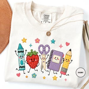 May include: An ivory t-shirt featuring a graphic print of a smiling blue crayon, a red strawberry, purple scissors, a purple spiral notebook, and a yellow pencil, surrounded by small colourful stars, hearts, and flowers. The label reads Comfort Colors.