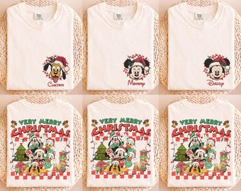 Può includere: Una collezione di magliette color bianco sporco con grafiche di personaggi Disney. Alcune magliette mostrano teste di personaggi come Pluto, Minnie o Mickey con testo sotto, mentre altre mostrano una scena di gruppo con il testo Very Merry Christmas.
