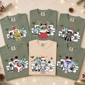 May include: Five sage green and one cream t-shirt featuring Star Wars character graphics with Christmas themes. Each shirt displays the Star Wars logo, holiday icons like Santa hats, holly, and ornaments, and characters including C-3PO, Grogu, Darth Vader, BB-8, a Stormtrooper, and R2-D2.