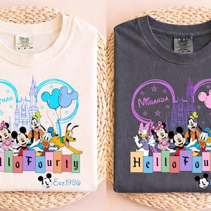 May include: Two t-shirts, one cream and one charcoal, featuring a Disney-themed design. The design includes Mickey Mouse and friends, the text "Hello Fourty", and the name "Jonathan" or "Miranda". The shirts have a crew neck.