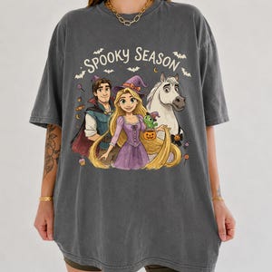 May include: A charcoal gray short-sleeve t-shirt featuring a graphic print of a man, a woman with long blonde hair wearing a witch hat, a white horse, and a small green lizard. The text SPOOKY SEASON is printed above with bats and candy.
