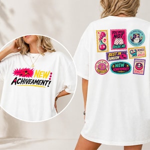 May include: White t-shirt with the text "NEW ACHIEVEMENT!" in a pink and yellow burst on the front. The back of the shirt features colorful stamp-like designs with phrases like "YOU WILL NOT BREAK ME" and "NEW ACHIEVEMENT!"
