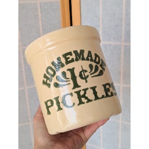 May include: A cream-colored ceramic crock with the words "HOMEMADE PICKLES" in green lettering. The crock has a slightly tapered shape and a wide opening. The text includes a stylized "1¢" symbol. The background is a light blue and white tiled wall.