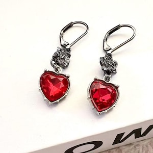 May include: A pair of dangle earrings featuring heart-shaped red gemstones. The earrings have a silver-colored metal setting and a floral design above the heart. The earrings are displayed on a white surface.