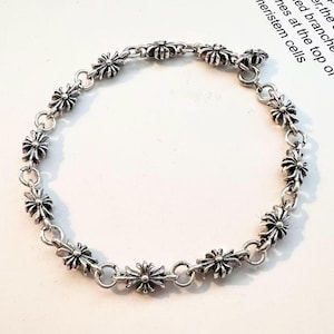 May include: Silver bracelet with a repeating floral pattern. The bracelet is made up of small, detailed flower-shaped links joined by circular rings. It has a simple hook and eye clasp. The bracelet is shown on a white surface.