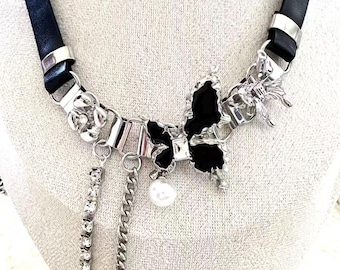 Puede incluir: Un collar gargantilla de cuero negro con detalles metálicos plateados. El collar presenta un colgante de lazo negro con una perla de imitación y detalles de cadenas colgantes. El diseño incluye una cadena de pedrería y una cadena de eslabones.
