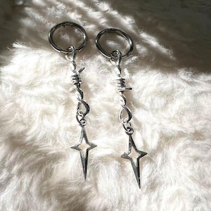 Pode incluir: Um par de brincos prateados com um design de arame farpado e um pendente em forma de estrela. Os brincos pendem de argolas circulares, sobre uma superfície branca e fofa.