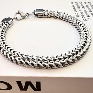 Może przedstawiać: Srebrna bransoletka z plecionym wzorem łańcucha. Bransoletka wykonana jest z błyszczącego metalu i posiada bezpieczne zapięcie. Bransoletka jest prezentowana na białej powierzchni. Widoczny jest tekst "NEWTON".