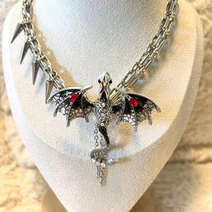 May include: A silver-toned necklace featuring a dragon pendant with black and red accents. The dragon has detailed wings and a serpentine tail. The necklace includes a chain with spike accents and is displayed on a white bust.