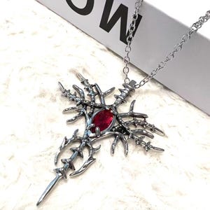 May include: Silver-toned pendant necklace featuring a cross design with a central red gemstone. The cross has a spiky, thorny appearance. The necklace is displayed on a white, textured surface.