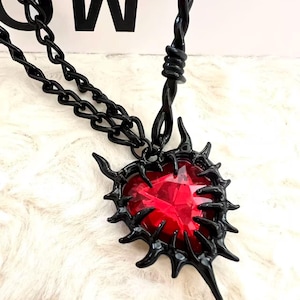 Può includere: Collana nera con un ciondolo a forma di cuore in pietra rossa. Il ciondolo è incorniciato da un design nero appuntito. La collana ha una catena e un elemento attorcigliato vicino al ciondolo.