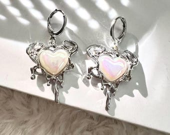 May include: A pair of silver earrings. Each earring has an iridescent heart-shaped centre stone, set within a decorative frame. The design includes a dangling element below the heart and a hoop closure.