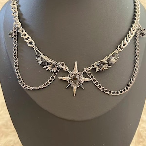 May include: Silver-toned necklace featuring a central starburst pendant with a dark stone. The necklace has chain detailing and small wing accents on either side, creating a gothic or celestial aesthetic.