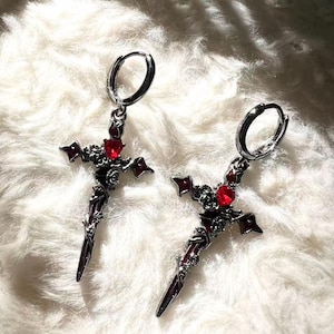 Puede incluir: Un par de pendientes de cruz plateados con detalles de piedras preciosas rojas. Los pendientes presentan un diseño en forma de daga con detalles intrincados y un cierre de aro. Las cruces están sobre un fondo blanco y esponjoso.