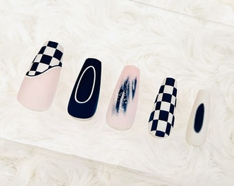 May include: A set of five false nails with different designs. One nail has a black and white chequered pattern, others are navy blue, pink with blue accents, and a white oval design. The nails are rectangular in shape.