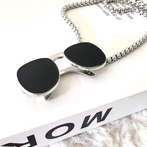Puede incluir: Un collar plateado con un colgante con forma de gafas de sol. Las gafas de sol tienen lentes negras y una montura plateada. El collar está sobre una superficie blanca con la palabra "MORE" visible.
